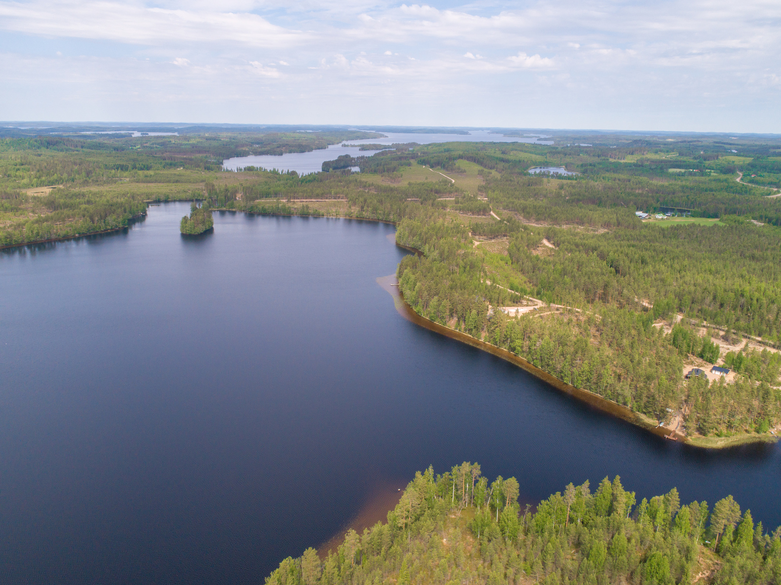 Kesämaisemassa metsäinen kannas erottaa kaksi vesialuetta. Rannoilla on mökkejä. Ilmakuva.
