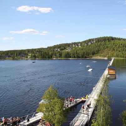 Järven ylittävällä kävelysillalla on ihmisiä. Vastarannan rinteessä mökkejä. Ilmakuva.