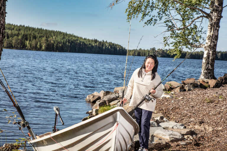 Nainen kalastusvälineiden kanssa sähköperämoottorilla varustetun veneen vieressä järven rannalla.