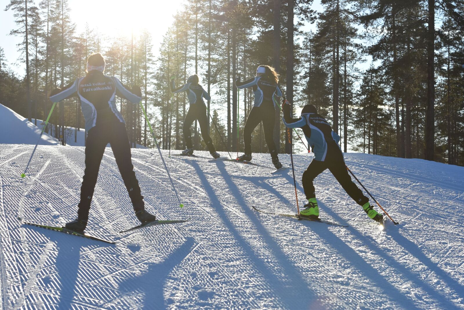 Neljä hiihtäjää nousee vauhdilla mäkeä ylös aurinkoisena talvipäivnä.