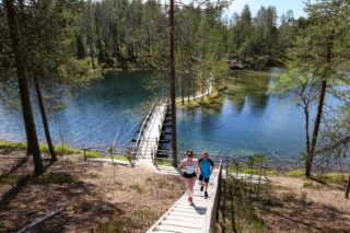 Kaksi henkilöä nousee portaita, jotka nousevat järven ylittävältä sillalta. Kesäinen maisema.