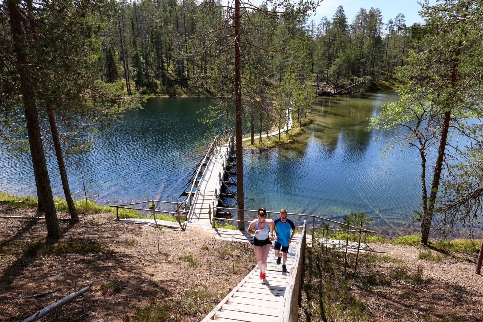 Kaksi henkilöä nousee portaita, jotka nousevat järven ylittävältä sillalta. Kesäinen maisema.