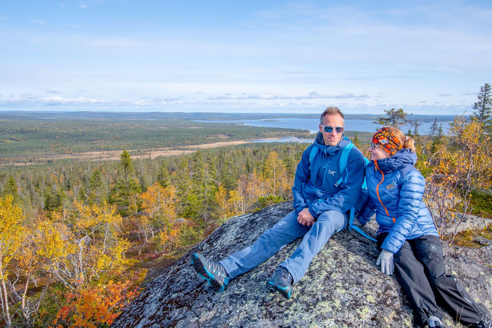Kaksi henkilö istuu ison lohkareen päällä vaaran laella. Lehtipuut ovat ruskan väreissä.