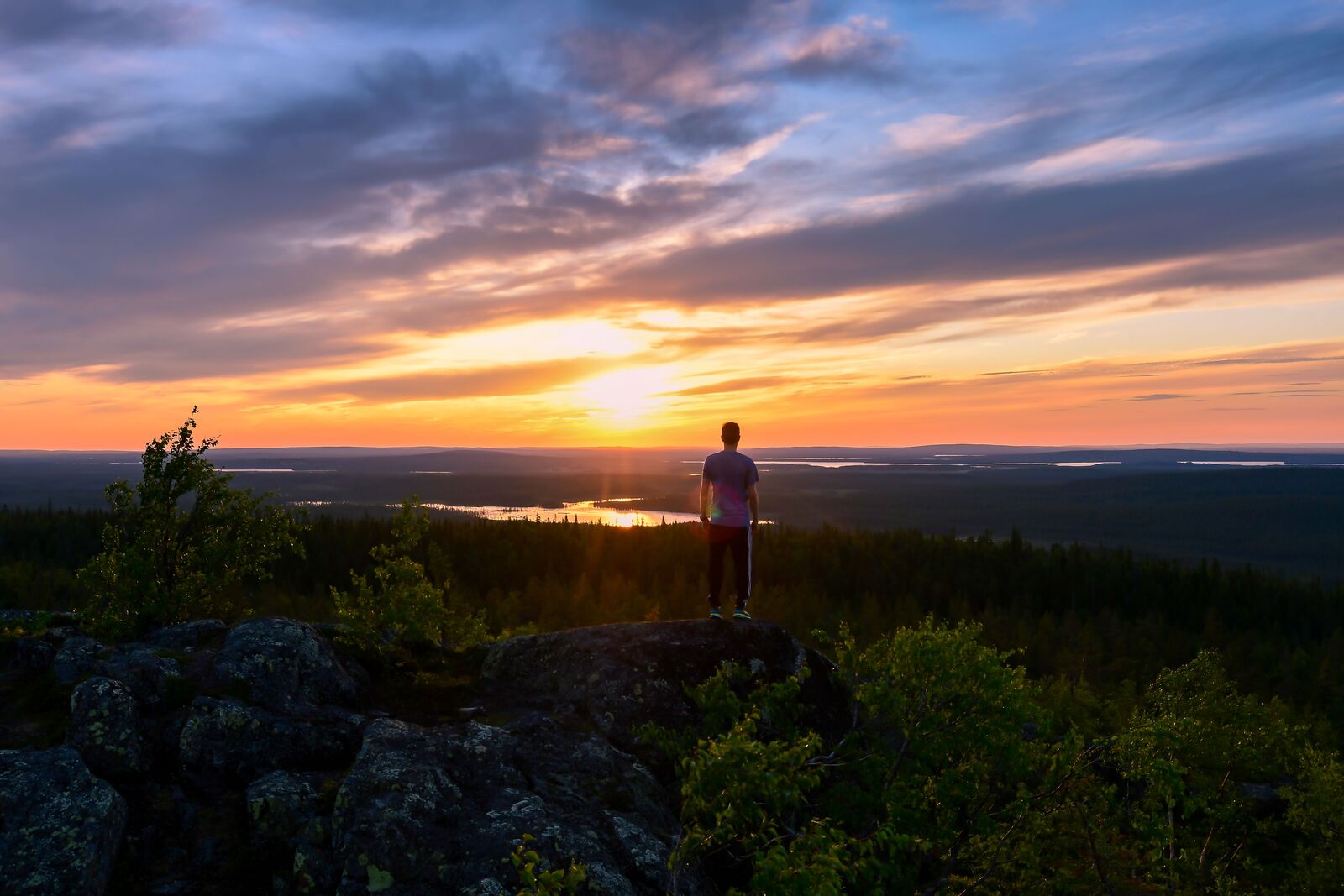 Henkilö seisoo tunturin laella katsellen kesäistä värikästä auringonlaskua.