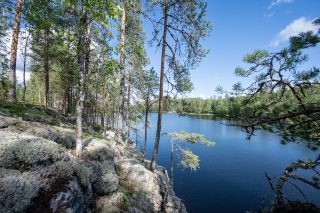 Rantakalliolle paistaa aurinko, vastarannalla näkyy metsää.