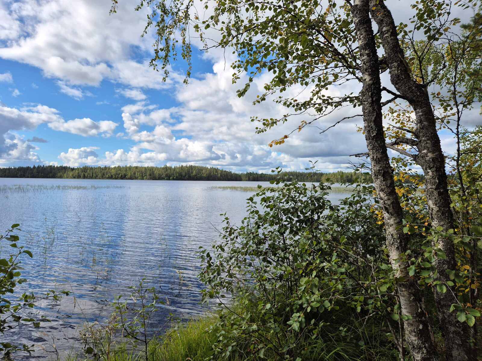 Maisema järvelle, aurinko paistaa, taivaalla on pilviä, vastaranta näkyy kaukaisuudessa.