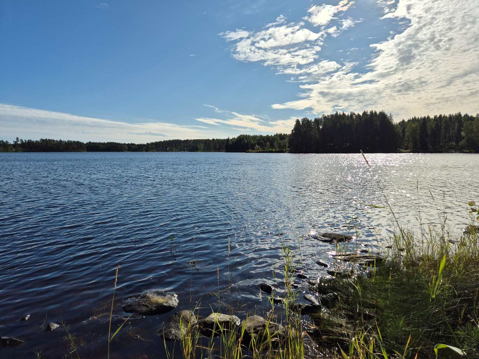 Järven pinta aaltoilee tuulessa, aurinko paistaa, rantavedessä on kiviä.