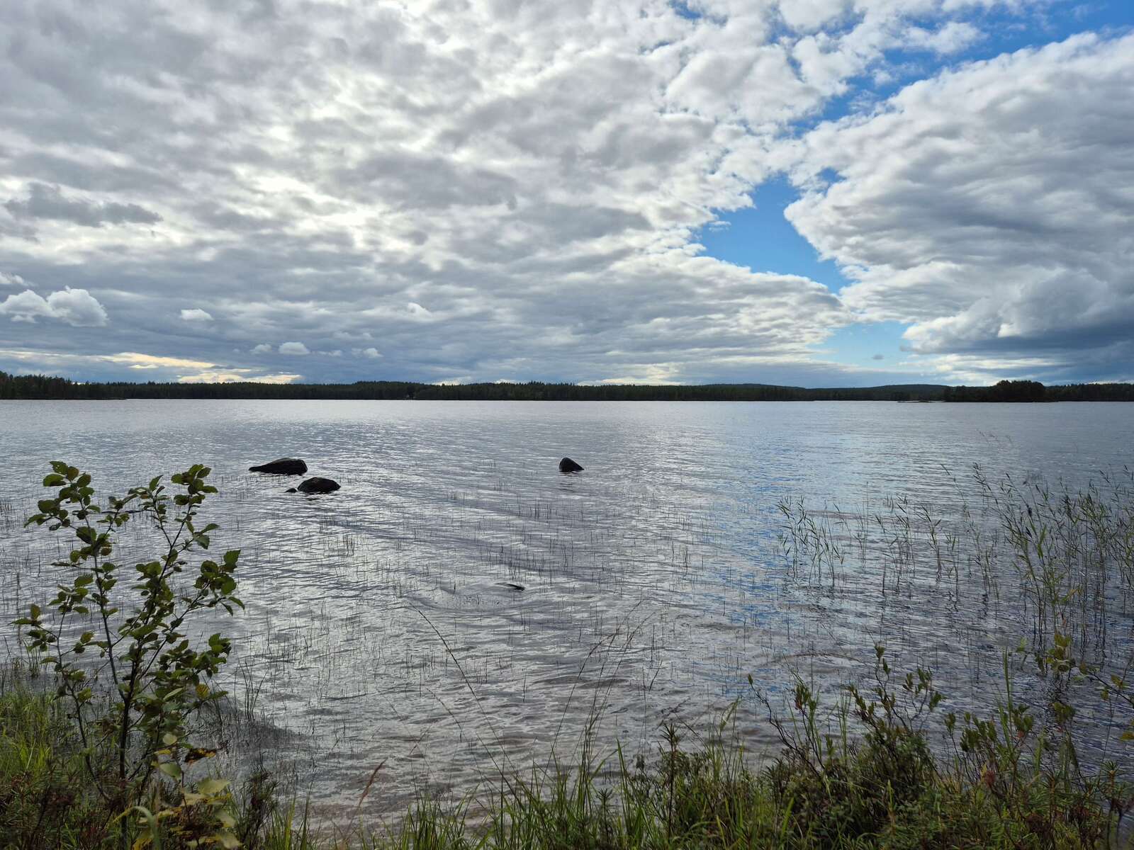 Maisema järvelle, vastaranta näkyy kaukana mustana nauhana, järvestä pilkistää isoja kiviä.