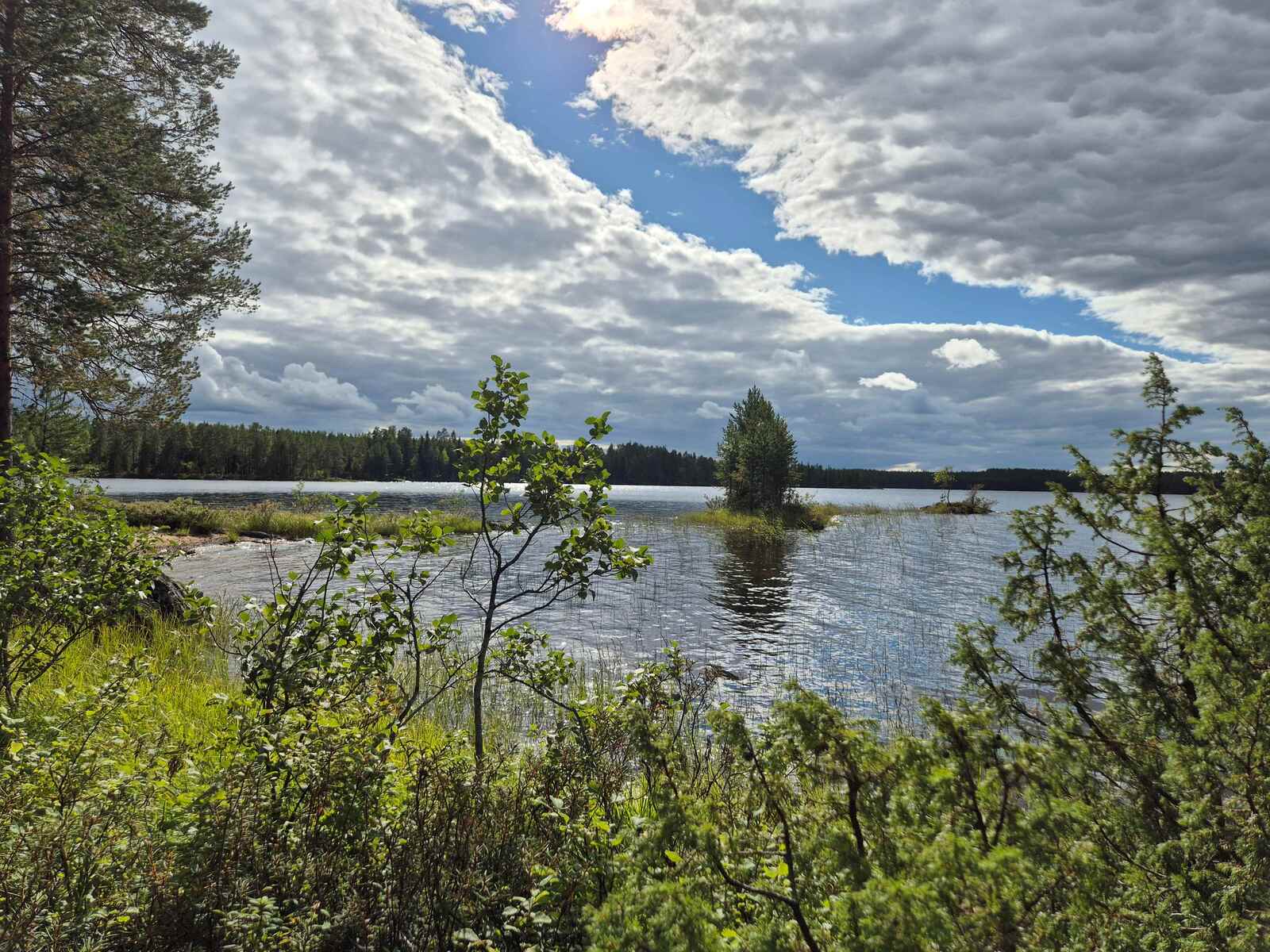 Järvimaisemassa on rannan edustalla pieni saari, taivaan pilviverhossa on yksi repeämä.