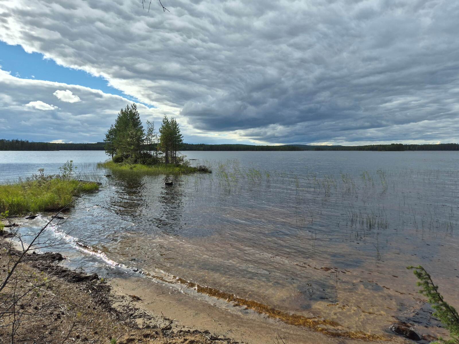 Maisema järvelle, luonnonhiekkarantaan lyö aalto, rannan läheisyydessä on pieni saari.