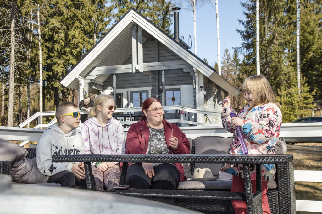 Äiti ja kolme lasta istuvat puutarhasohvalla harmaan saunamökin edustalla.
